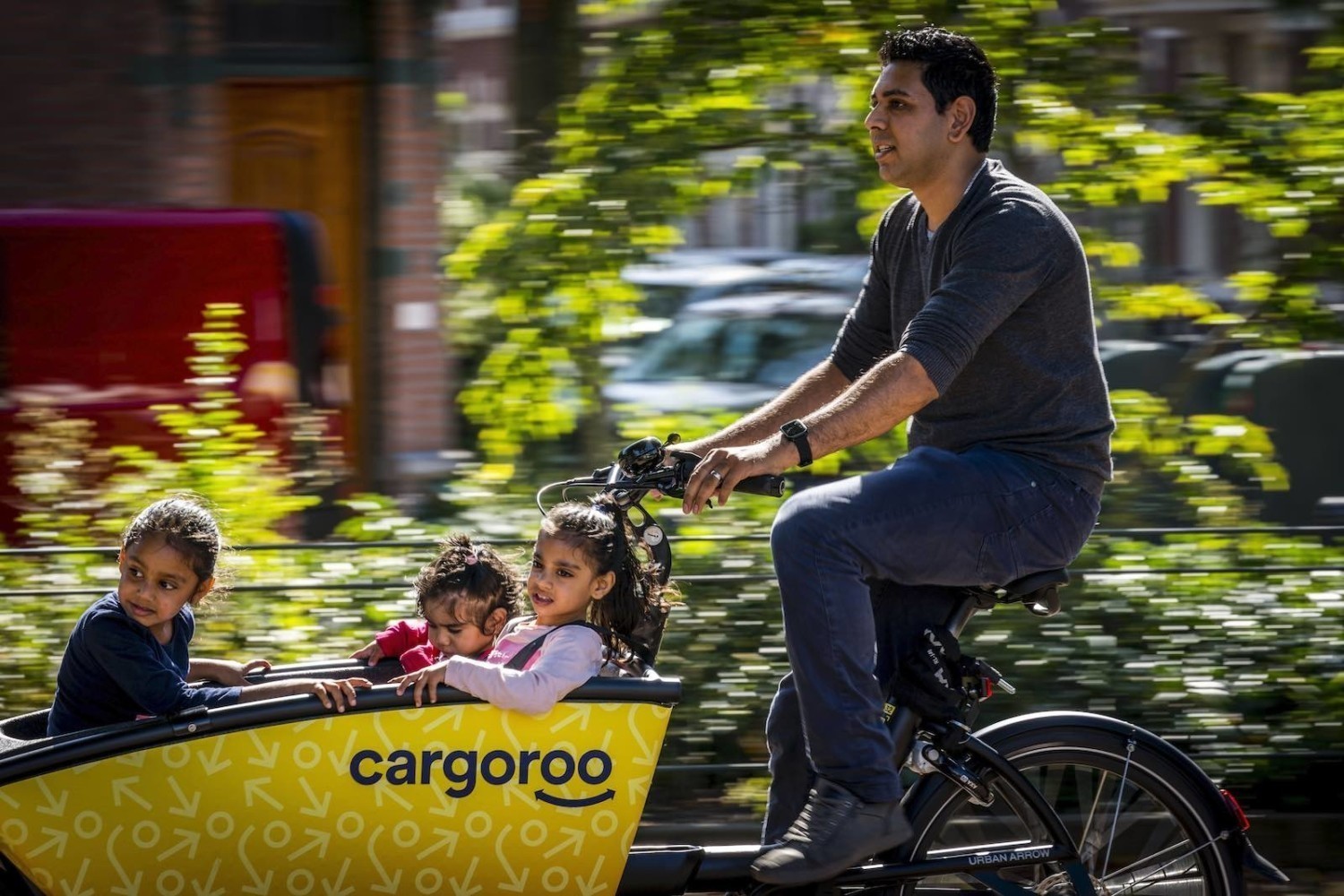 A parent rides three children through the city in a Cargoroo cargo bike